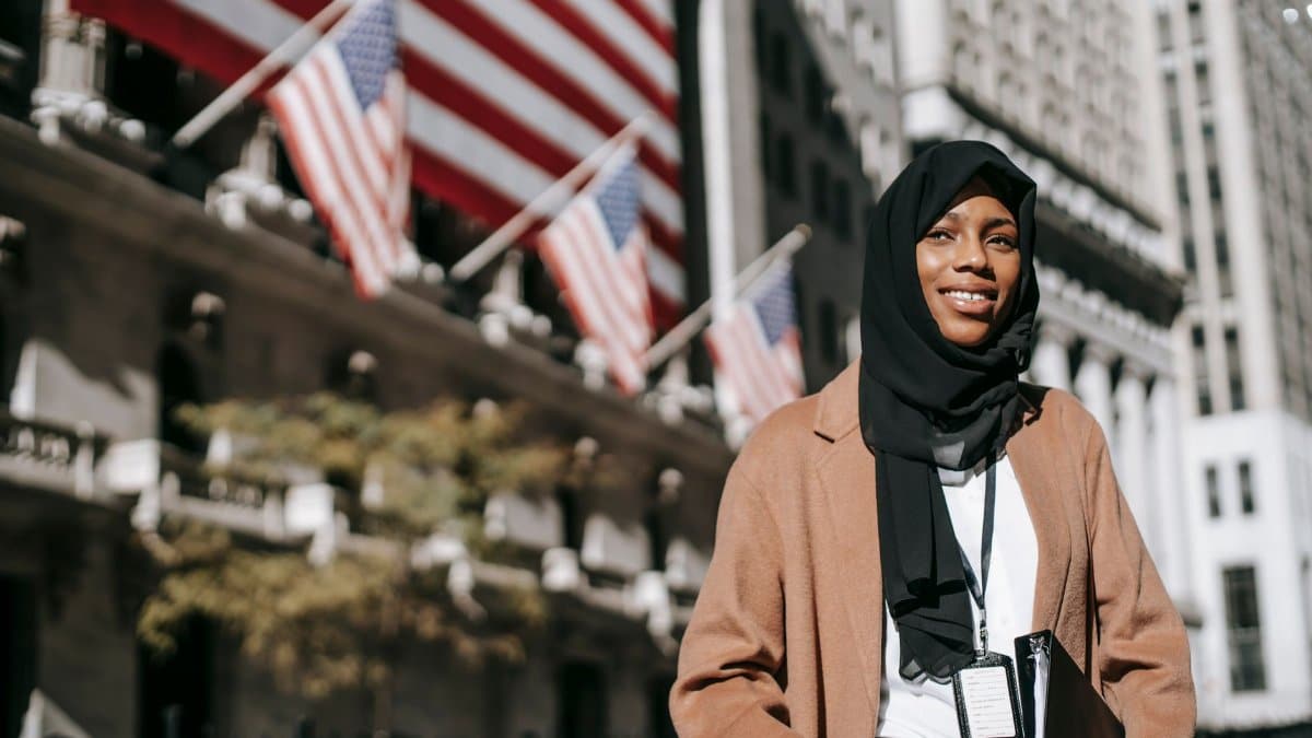 From below of cheerful African American female ambassador with folder wearing hijab and id card looking away while standing near building with American flags on blurred background