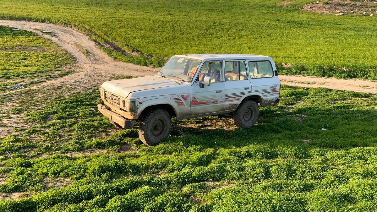 Old SUV on a dirt path in a lush green field with mountain backdrop.