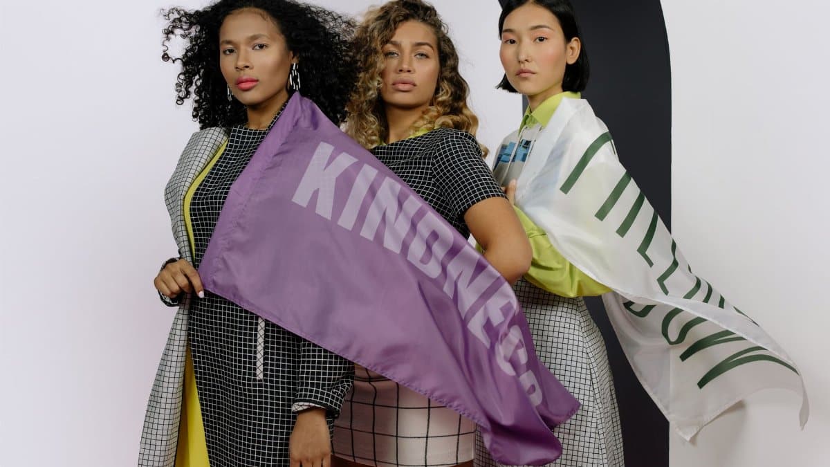 Three women stand united, celebrating diversity with kindness-themed flags.