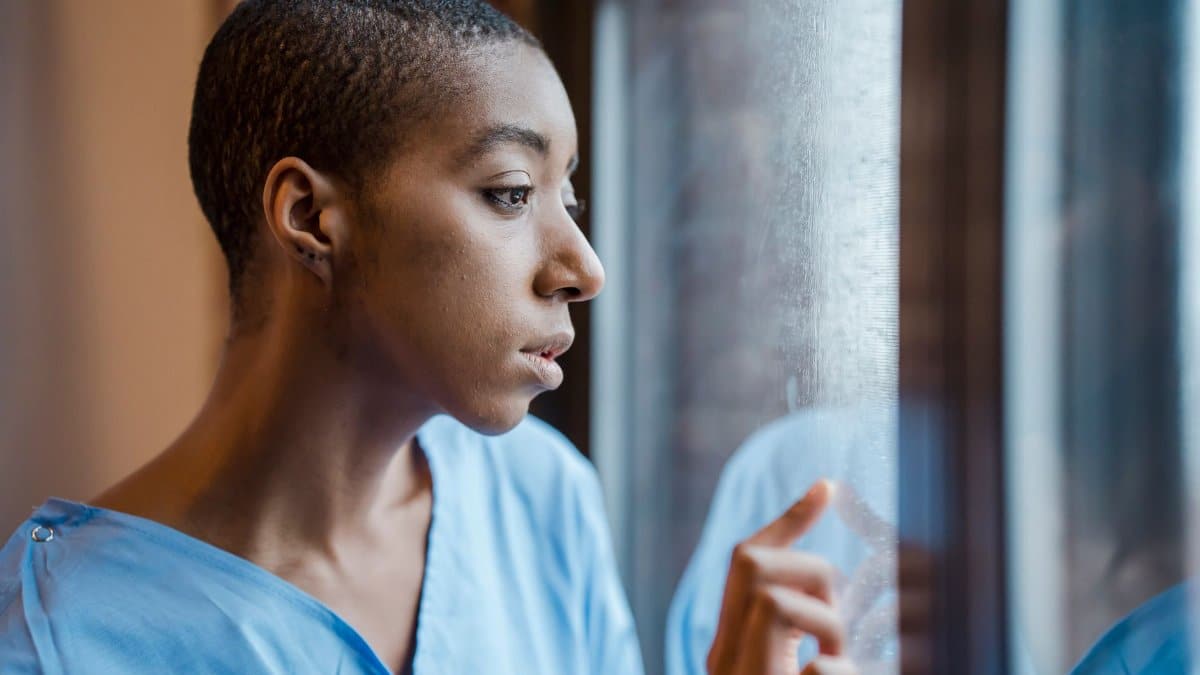 Upset African American female patient in medical wear touching window while looking away with concern