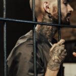 A tattooed man behind prison bars, contemplating life inside a penitentiary.