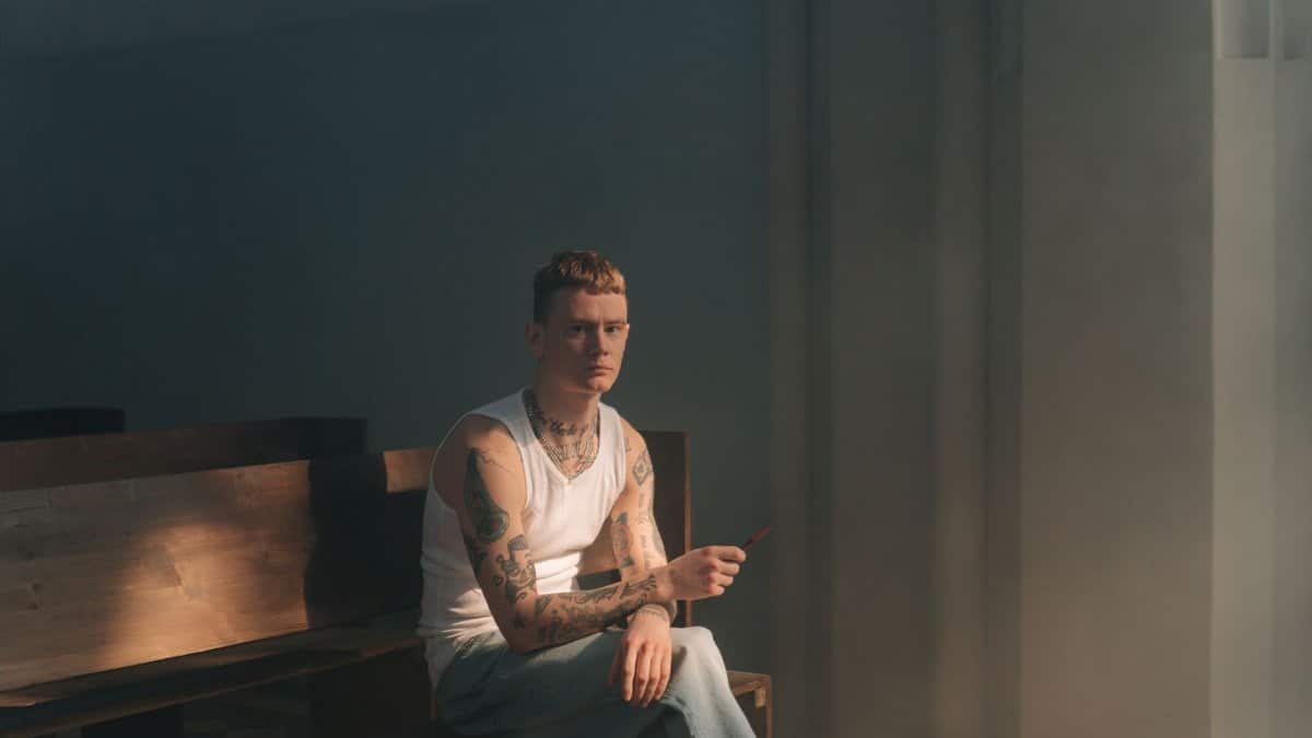 A tattooed man sits in a church pew, wearing a white tank top, reflecting.