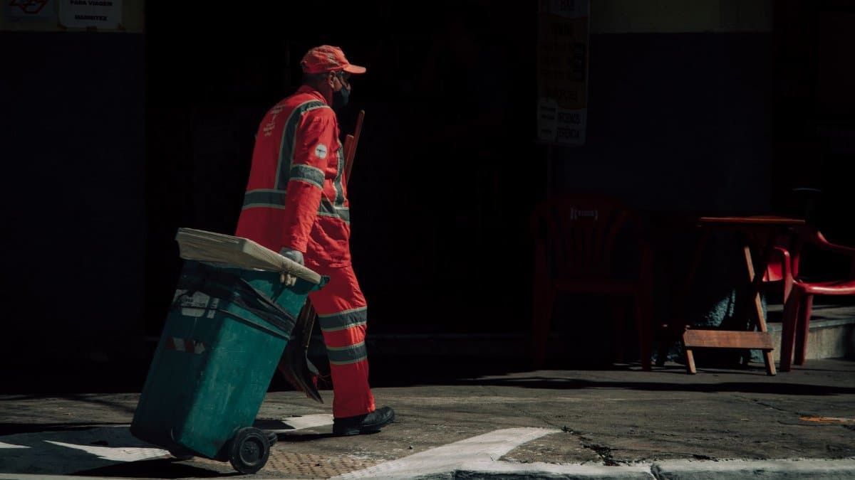 Municipal worker in uniform pulling a trash bin on an urban street.