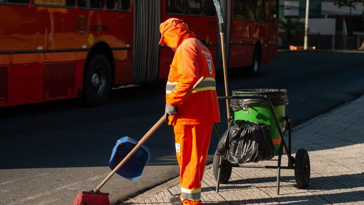 Street cleaner in bright orange uniform sweeping sidewalk near public bus in city setting.