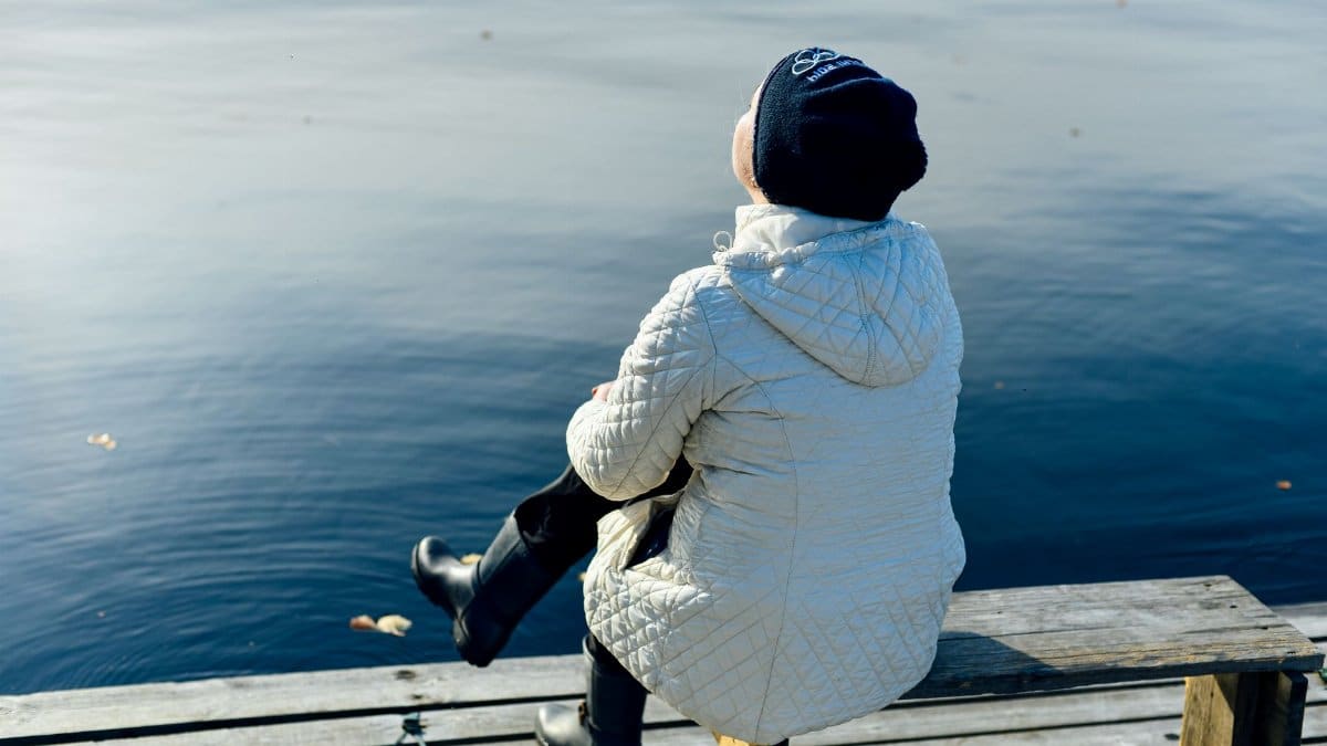 A person sits on a wooden dock, wearing a bubble jacket and a knit hat, gazing at the water.