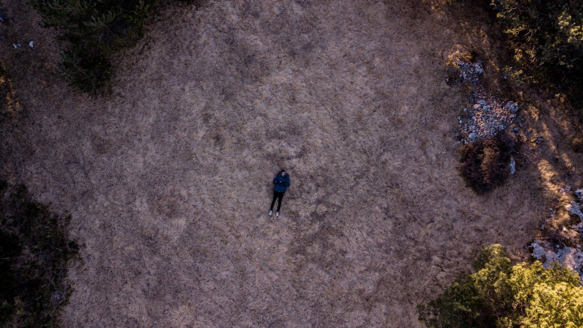 A solitary figure lies in a vast open field surrounded by nature, captured from above.