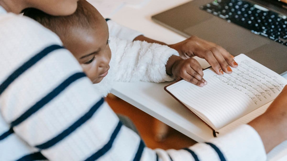 Ethnic baby with mother wearing casual outfit while sitting at table with opened notebook near laptop in bright room