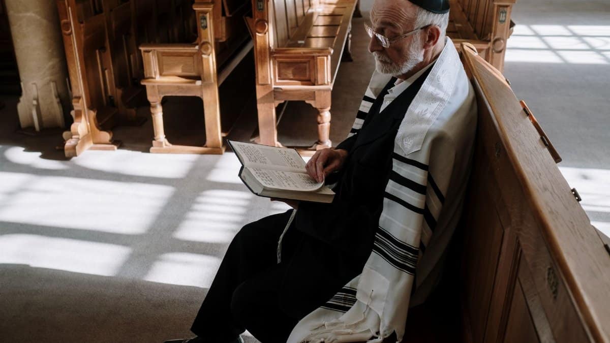 Elderly man wearing kippah and tallit, reading in a synagogue. Traditional Jewish setting.