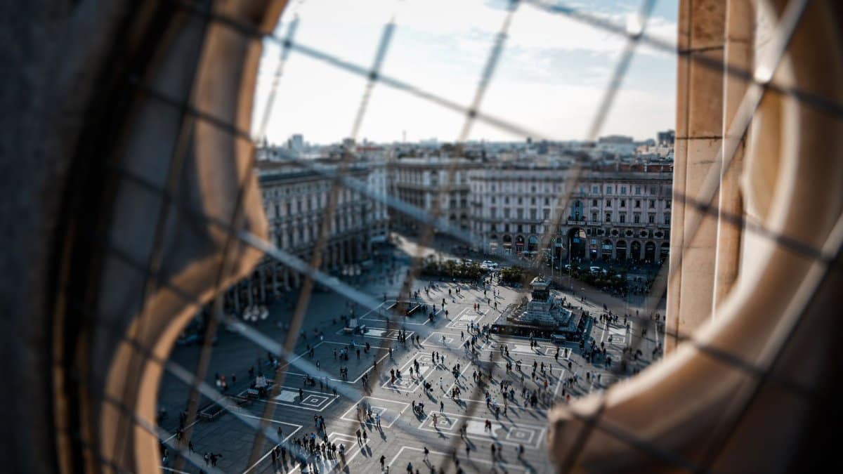 Crowd moving through Piazza del Duomo in Milan, Italy, seen from high above through ornate architectural details.