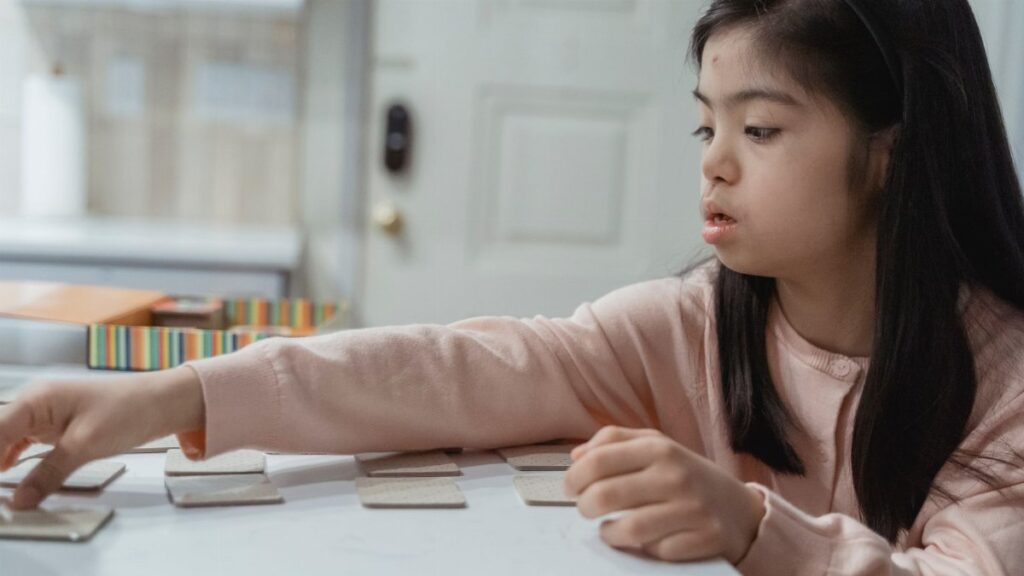 Young Asian girl with Down Syndrome engaged in a game indoors, wearing a pink sweater.