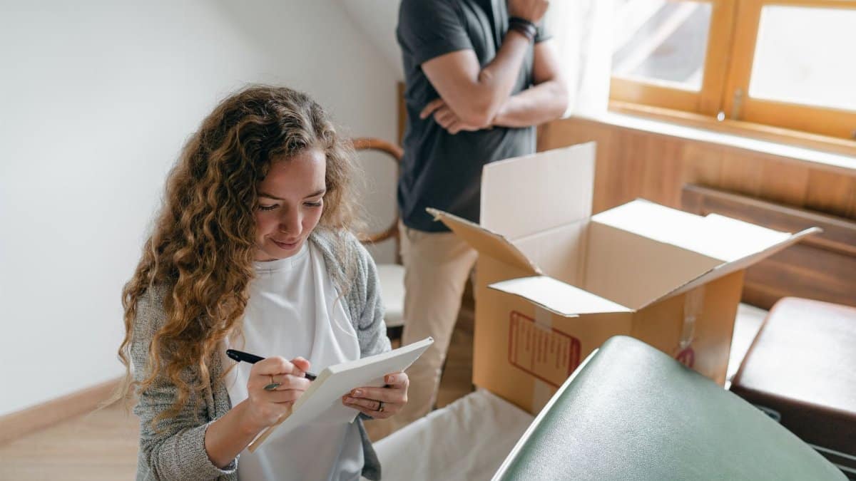 Pensive couple taking notes in bedroom near window while packing stuff in carton box to move into new apartment