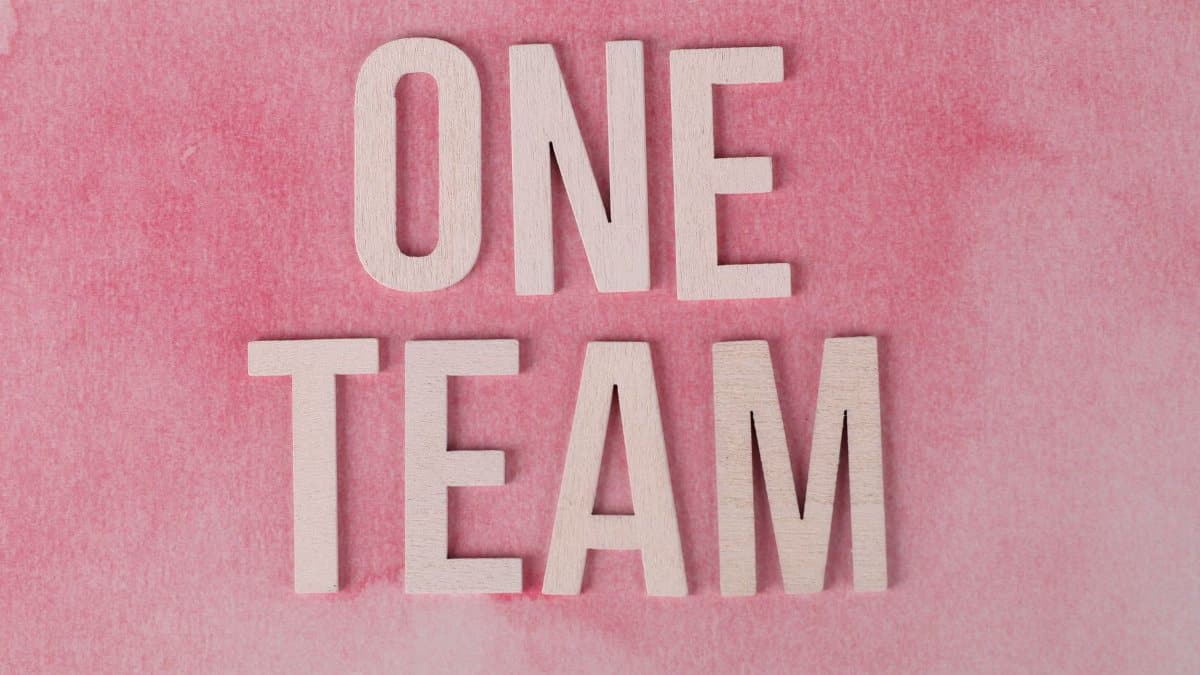 Wooden letters spelling 'One Team' on a pink textured background symbolizing unity.