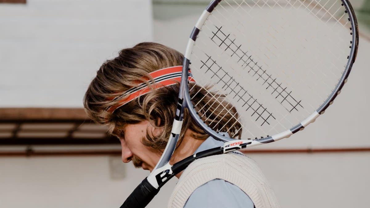 Tennis player with long hair wearing a sweatband holds racket on shoulder outdoors.