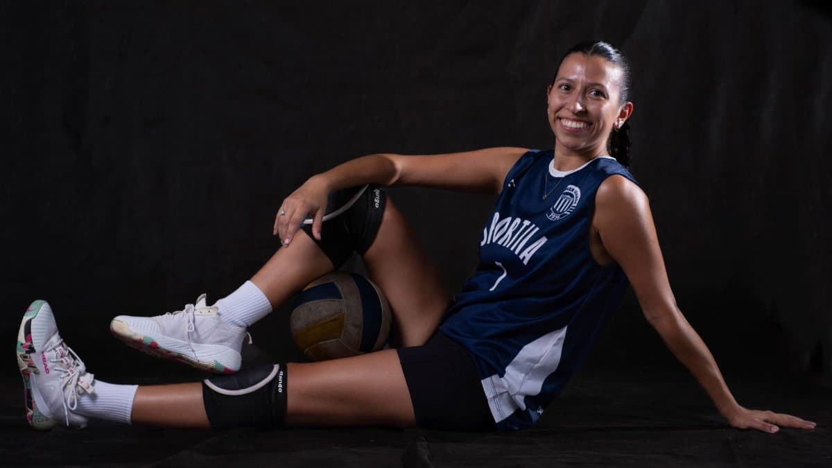 Smiling female volleyball player seated with ball, wearing knee pads, jersey, and sneakers.