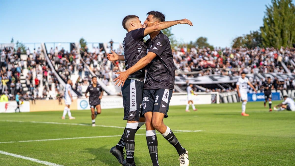 Two soccer players in black jerseys celebrate a goal on a sunny day at a packed stadium.