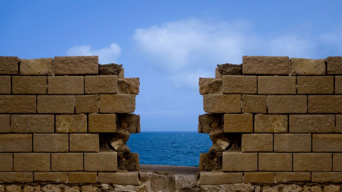 A stone wall with a gap overlooks the blue Mediterranean Sea under a clear sky.