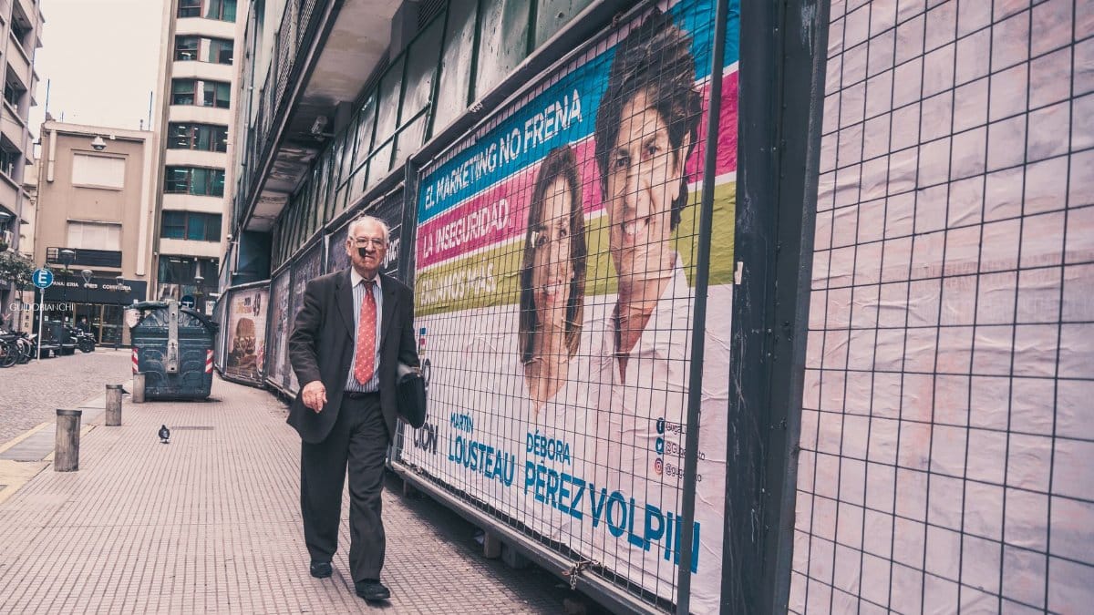 Elderly man walks past colorful political posters in an urban alleyway.