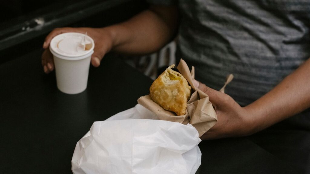 Unrecognizable male sitting at table in street cafe with cup of hot beverage and eating fried pie during lunch break