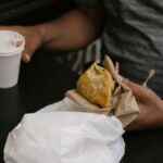 Unrecognizable male sitting at table in street cafe with cup of hot beverage and eating fried pie during lunch break
