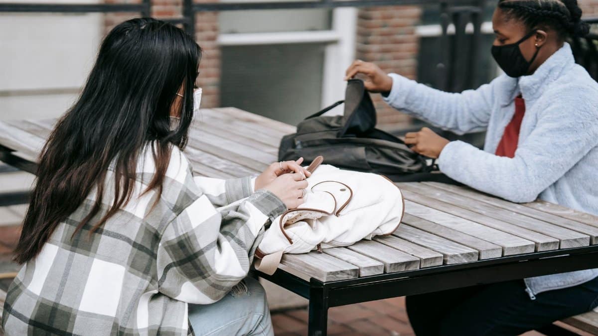 Young multiethnic female students in protective masks with backpacks sitting at wooden table in campus and preparing to work on task together