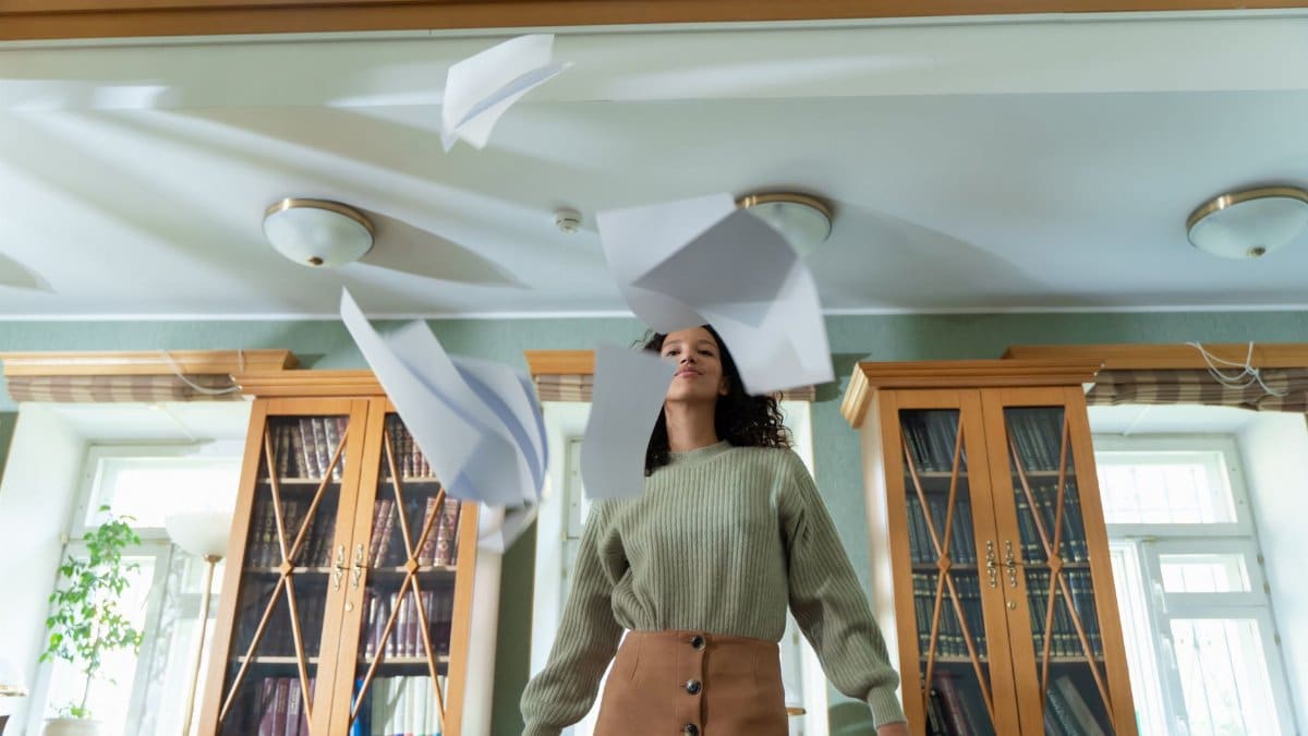 Confident woman throws papers in an academic library, showcasing freedom and academic rebellion.