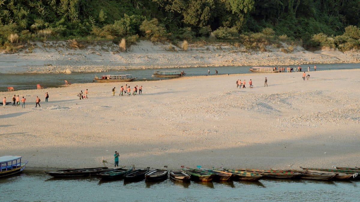 Scenic view of a riverbank with boats lined up and groups of people walking, set against a lush forest background.