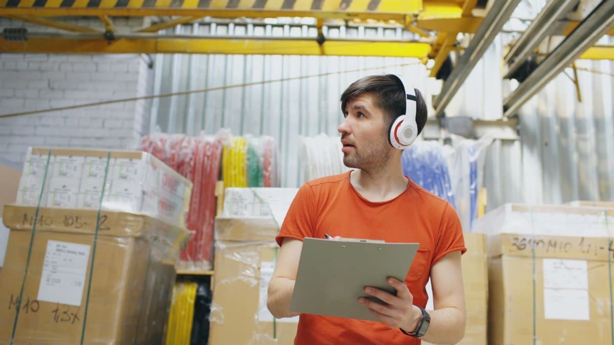 Young male warehouse worker with headphones checks inventory on clipboard in industrial setting.
