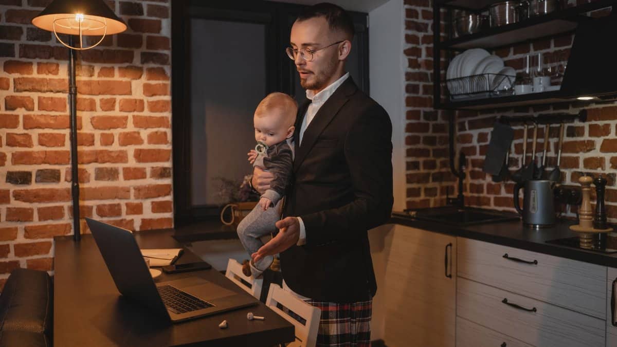 A father multitasks with a baby while working on his laptop in a cozy kitchen setting.