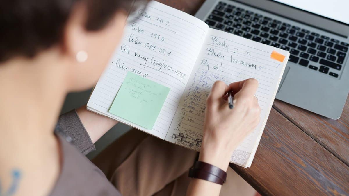 An adult writing in a notebook with a laptop on a wooden table for work or leisure.
