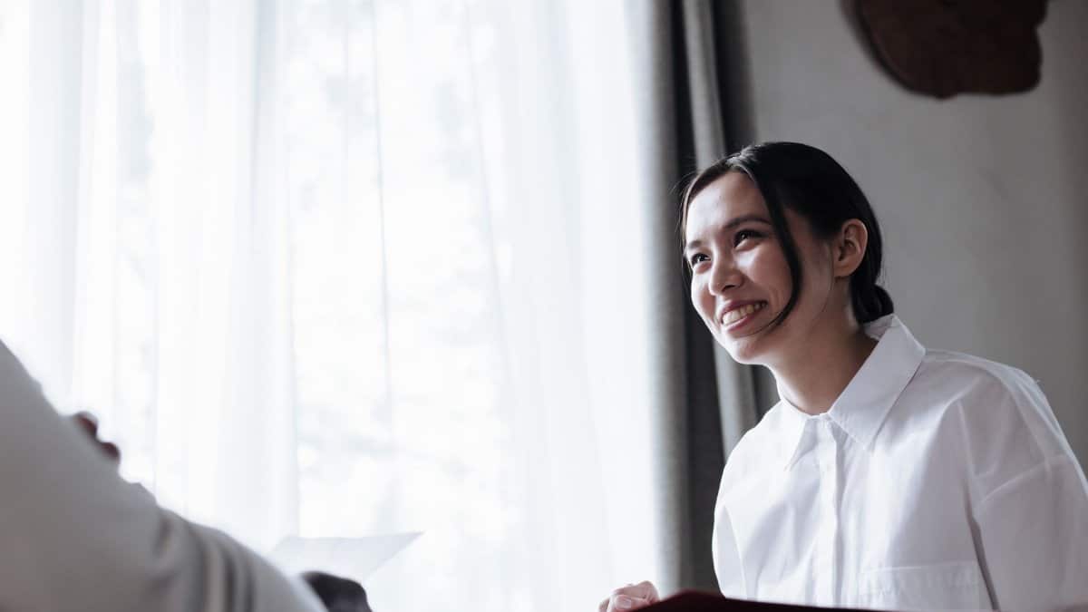 A smiling woman in white shirt engaged in conversation indoors by a window.