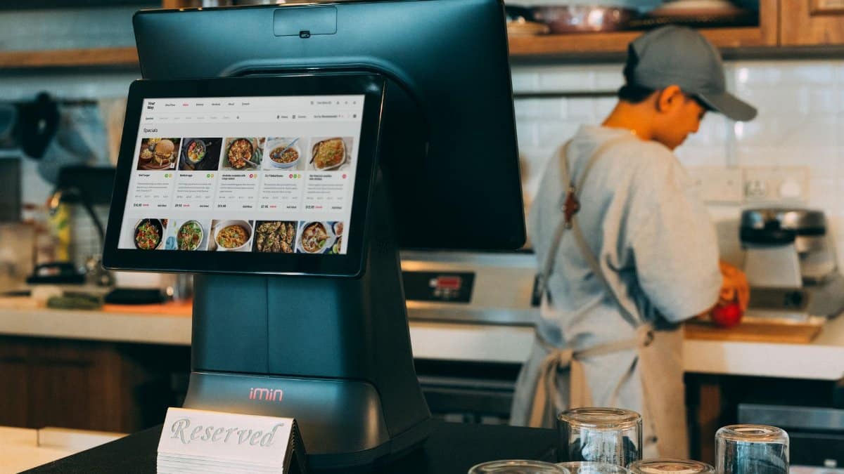 Digital ordering system in a restaurant kitchen with a touchscreen display and a chef at work.
