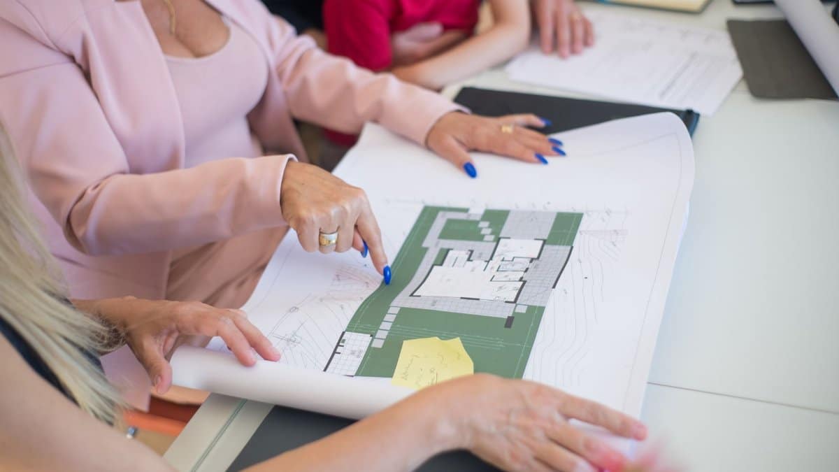 A real estate agent in a pink blazer shows floor plans to clients, discussing property details.