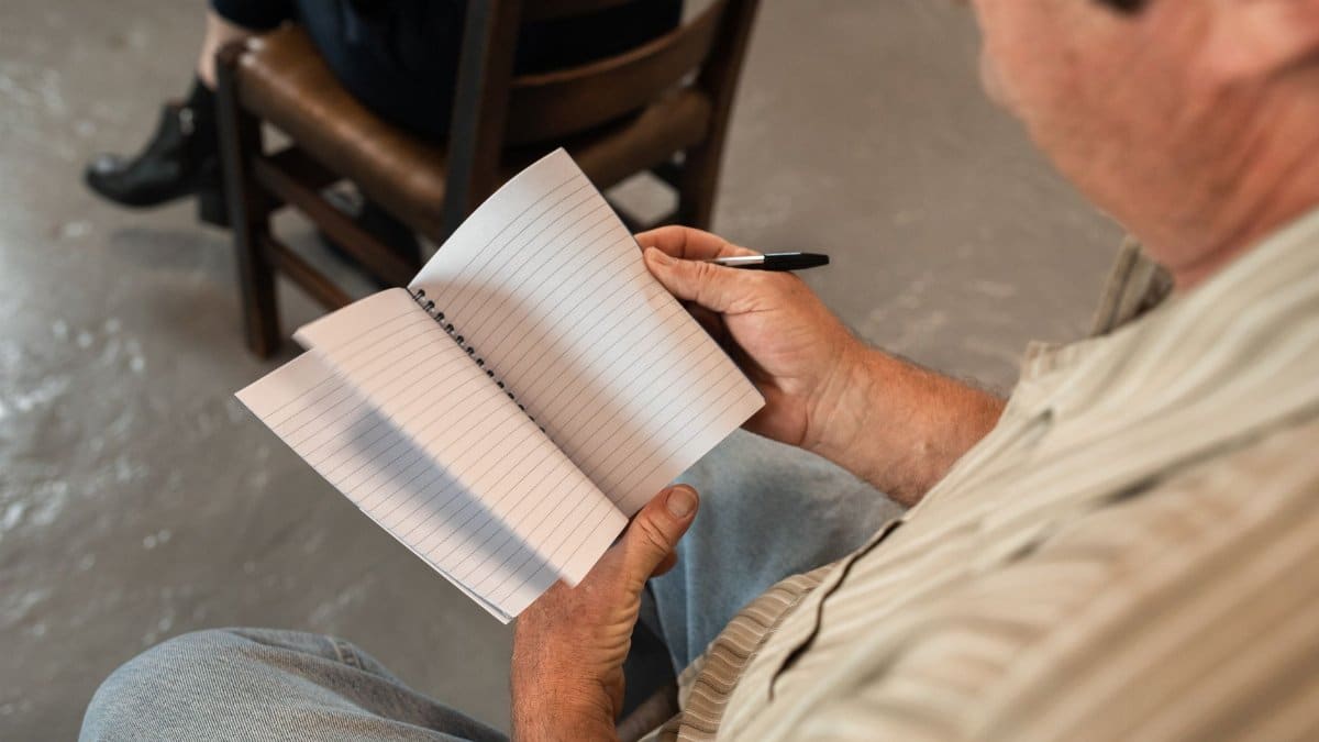 A man writing in a notebook during an indoor meeting or discussion.