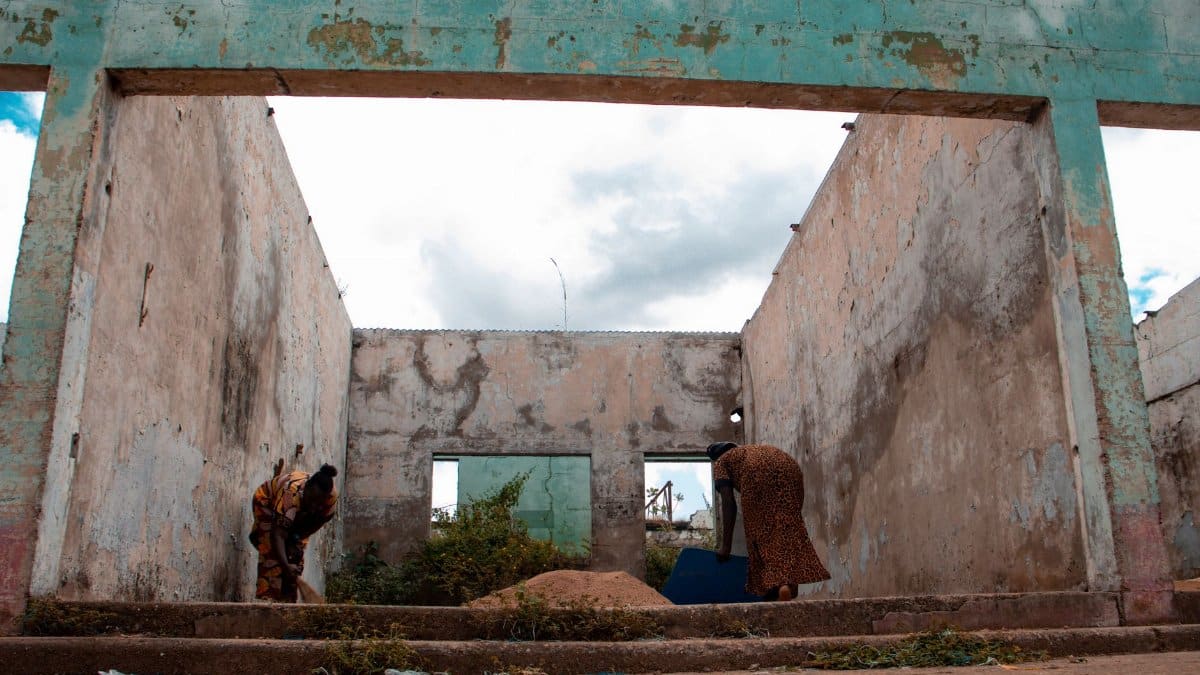 Two women cleaning rubble in a deserted, open-roof structure under cloudy skies.