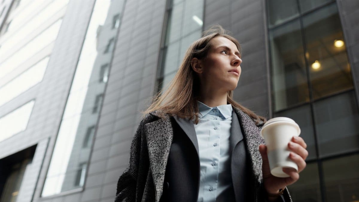Professional woman in a city street holding a coffee cup, gazing upwards.