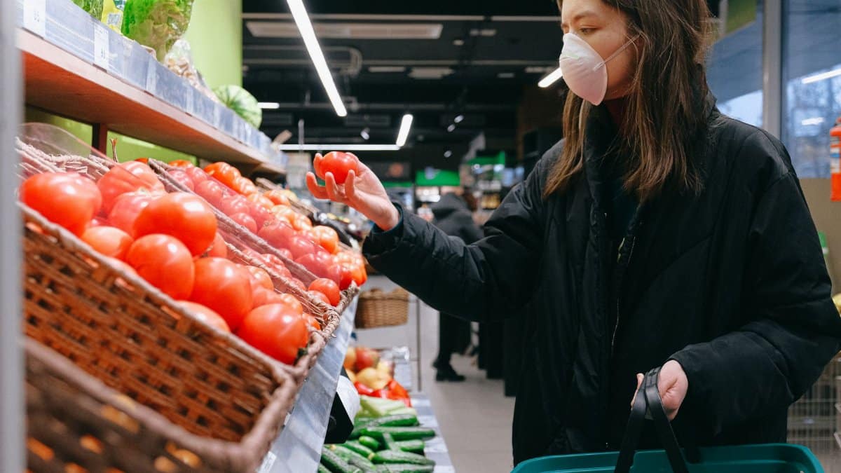 A woman in a face mask selects tomatoes at a grocery store, showcasing pandemic shopping habits.