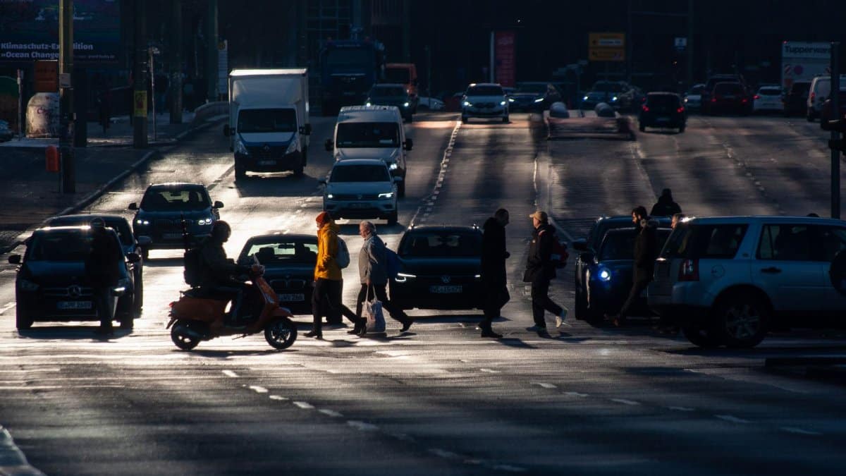 Morning traffic in an urban setting with pedestrians crossing a busy street amid cars and scooters.