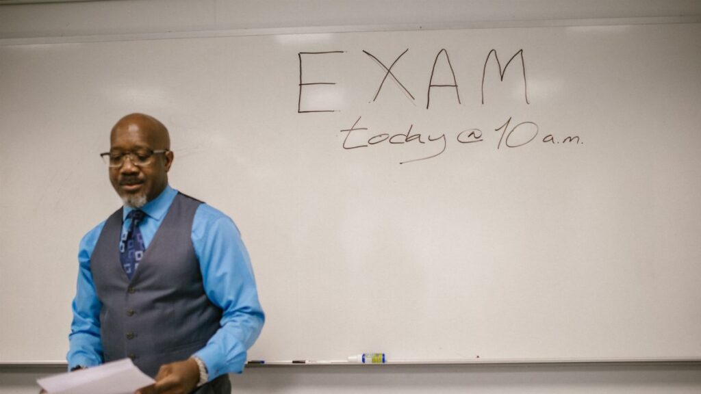 A professor stands next to a whiteboard with 'Exam today at 10 a.m.' written, indicating an upcoming exam in a classroom.