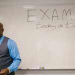 A professor stands next to a whiteboard with 'Exam today at 10 a.m.' written, indicating an upcoming exam in a classroom.