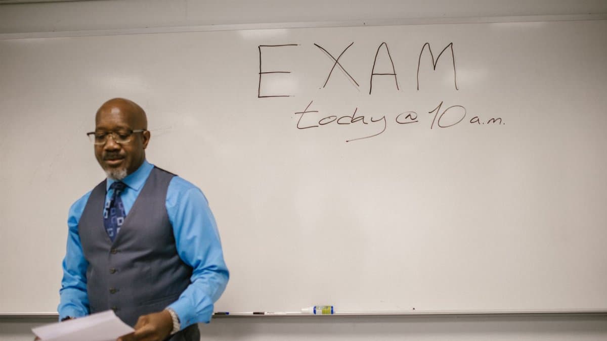 A professor stands next to a whiteboard with 'Exam today at 10 a.m.' written, indicating an upcoming exam in a classroom.
