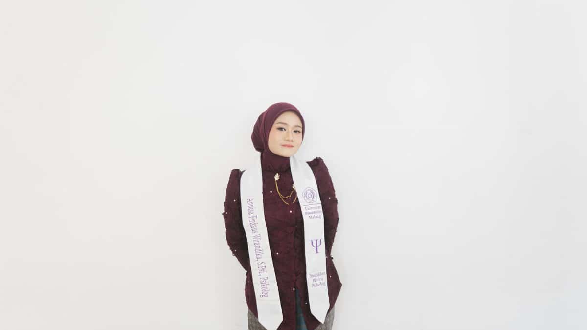 Young woman in graduation attire with maroon hijab and patterned outfit against a white background.