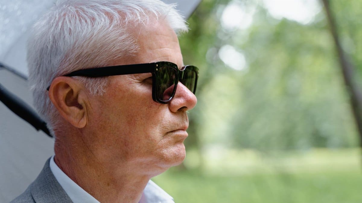 Profile view of a stylish senior man wearing sunglasses, standing outdoors with trees in the background.