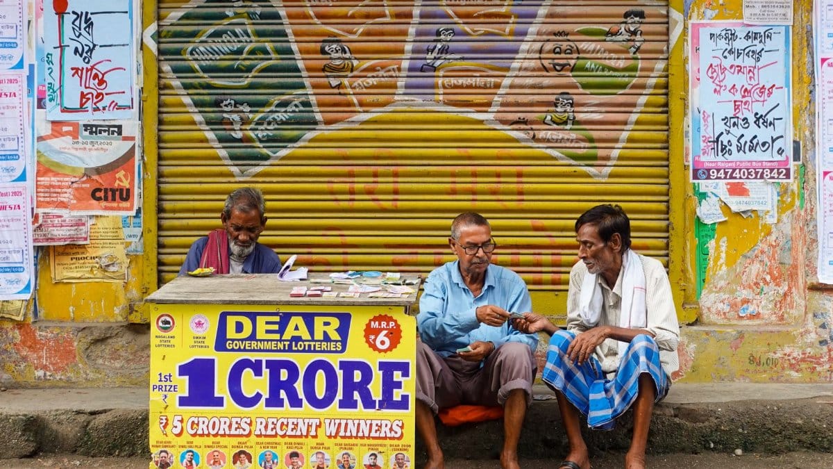 Three men sit at a lottery booth outside in Raiganj, India. Colorful posters and signs add vibrancy.