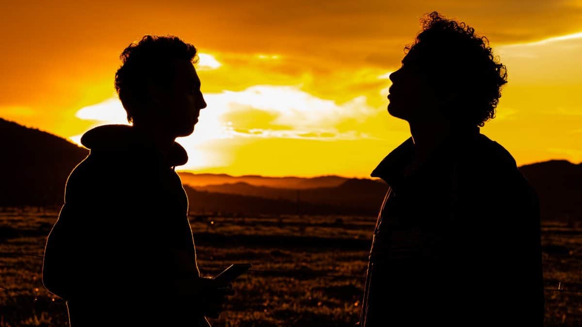 Two silhouetted figures stand against a vibrant sunset backdrop in a rural landscape.