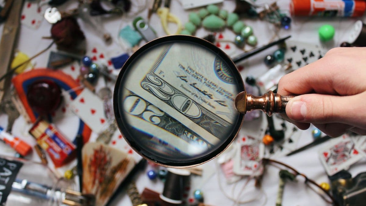 A hand holding a magnifying glass focusing on a twenty dollar bill amidst various scattered objects.