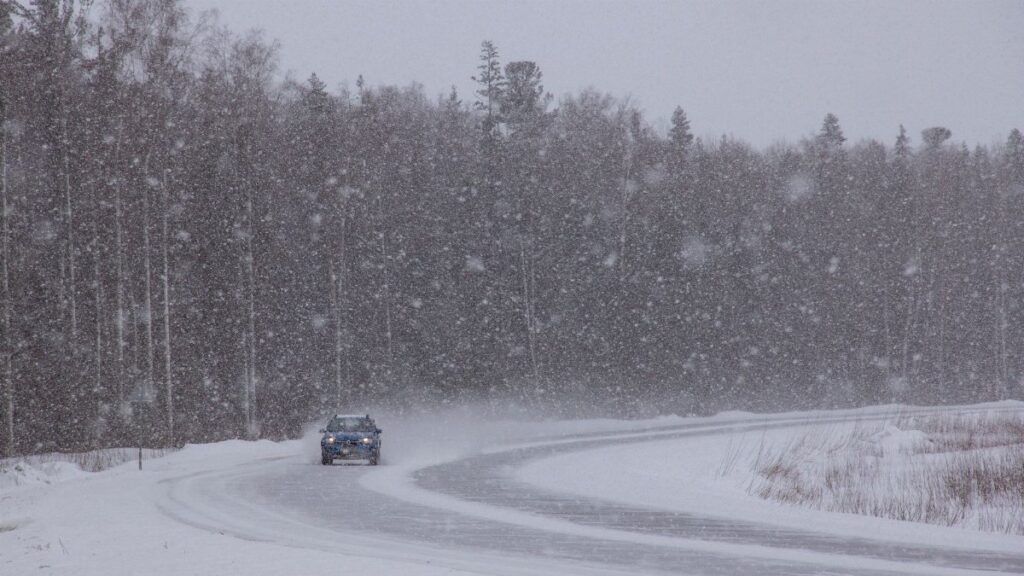 A car navigates a snowy forest road during a winter snowstorm, capturing the harsh weather conditions.