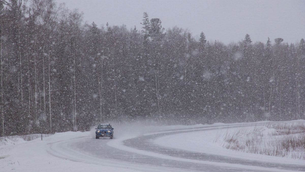 A car navigates a snowy forest road during a winter snowstorm, capturing the harsh weather conditions.