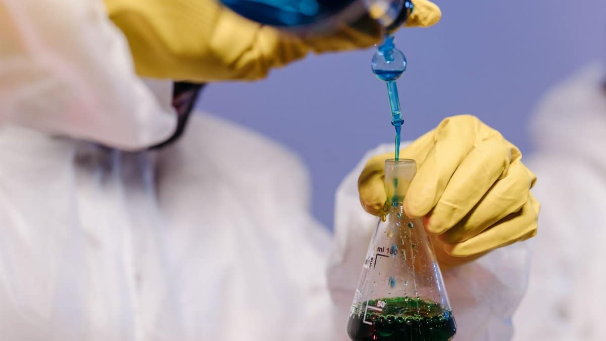 Scientist wearing gloves and lab coat mixing colored liquids in a flask.
