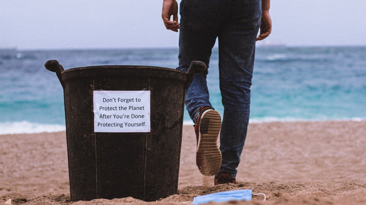 Man walking by beach trash can with a face mask and awareness message about protecting the planet.