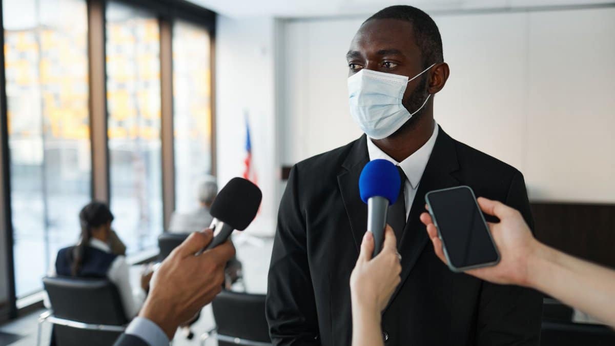 African American businessman in formal attire speaks to media while wearing a face mask in a conference room setting.